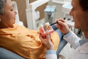 Closeup of male dentist holding a tooth model while explaining dental implant surgery to a new patient in a dental clinic.